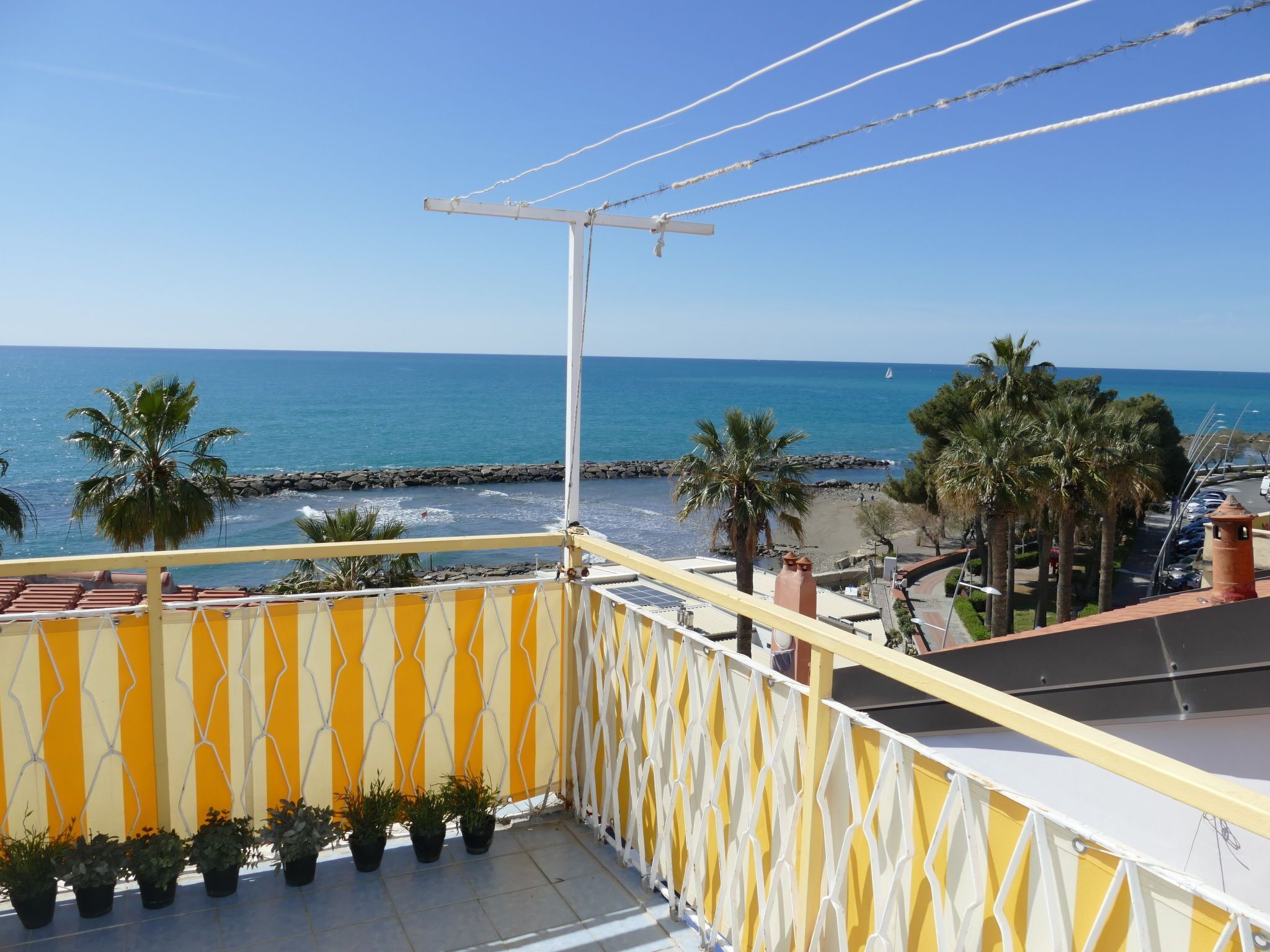 A balcony with a view of the sea, palm trees, and a rocky breakwater.