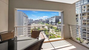 Un balcón con mesa y sillas, con vistas a un paisaje urbano con edificios modernos y vegetación.
