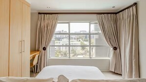 Habitación de hotel con cama, escritorio y vistas a la ciudad a través de amplias ventanas.