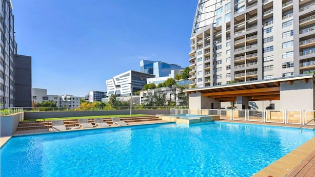 Una piscina con muelle de madera y sillones de descanso, rodeada de edificios modernos.