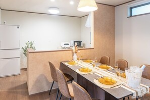 Ruokailutilassa on pitkä pöytä, johon on katettu kuudelle, taustalla näkyy keittiö ja oikean seinän puolella on ikkuna.