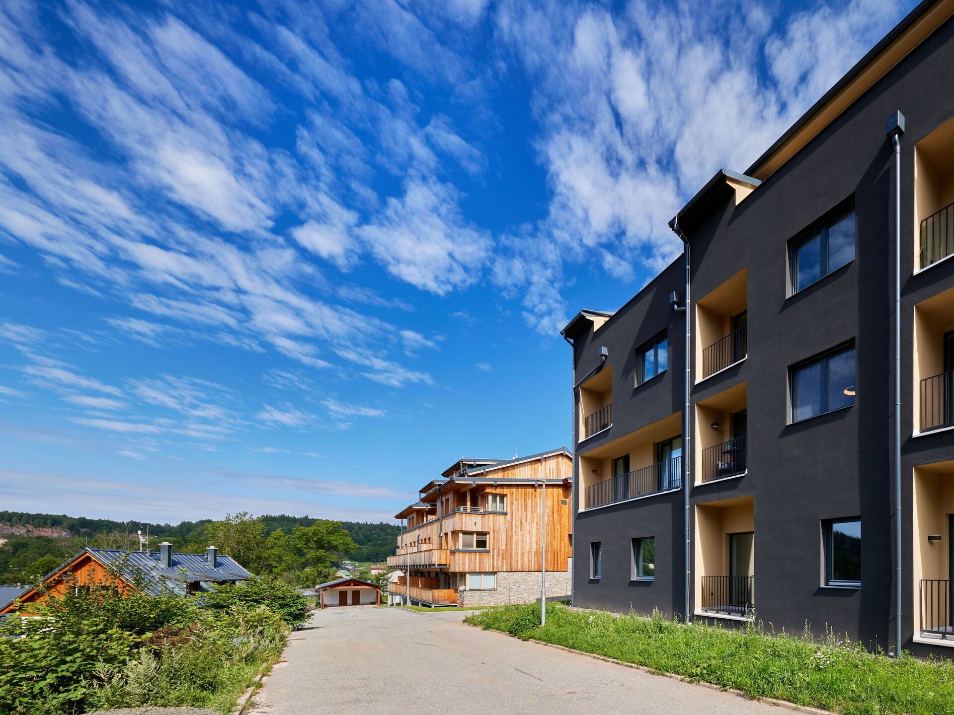 A modern multi-story building with balconies, a clear sky, and a road leading to a wooden building in the background.