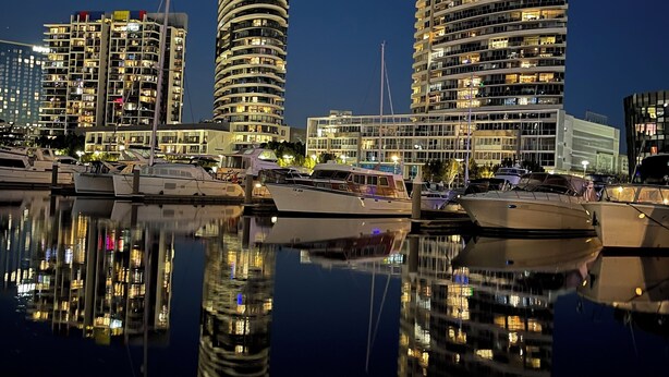 Een jachthaven aan het water met moderne gebouwen, waaronder een gebogen wolkenkrabber en een cilindervormige toren, die in het water worden weerspiegeld.