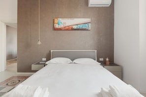 Una camera d'albergo moderna con un letto grande, un comodino, un condizionatore d'aria a parete e un quadro del ponte di Manhattan.