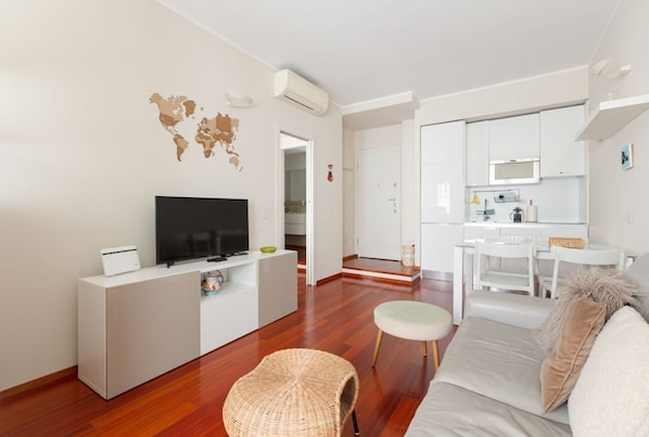 A modern living room with a flat-screen TV, a white console table, a wicker chair, a sofa, and a kitchen area in the background.