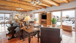 Ein geräumiges Wohnzimmer mit hohen Holzbalken, einem Kamin und großen Fenstern mit Blick auf verschneite Landschaft.