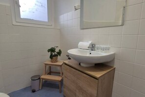 A bathroom with a white sink, a mirror, a small wooden stool, a trash bin, and a potted plant.