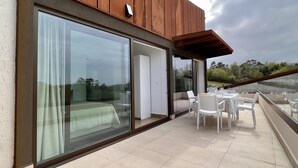 A modern building with large sliding glass doors, a covered outdoor seating area with white chairs, and a view of greenery in the background.