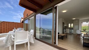 A modern outdoor dining area with a table and chairs, a sliding glass door leading to a kitchen, and a view of the surrounding landscape.