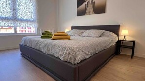 A bedroom with a large bed, a window with patterned curtains, a bedside table with a lamp, and a painting on the wall. - Cozy apartmet in Hoznayo (Entrambasaguas)