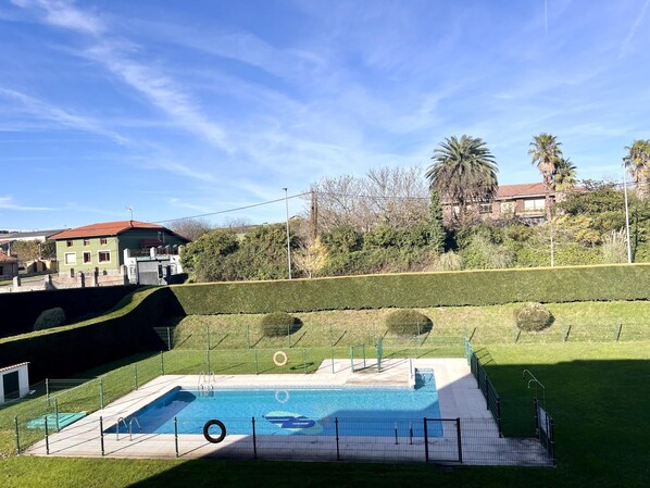 A swimming pool with a fence, a grassy area, and a building with a red roof in the background.