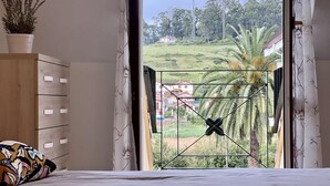 Un dormitorio con cama, cómoda de madera, planta en maceta y vista a un paisaje verde a través de una ventana con reja de metal.