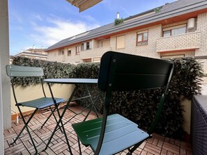 Um pátio com duas cadeiras verdes e uma mesa, piso de tijolos e uma sebe ao fundo.