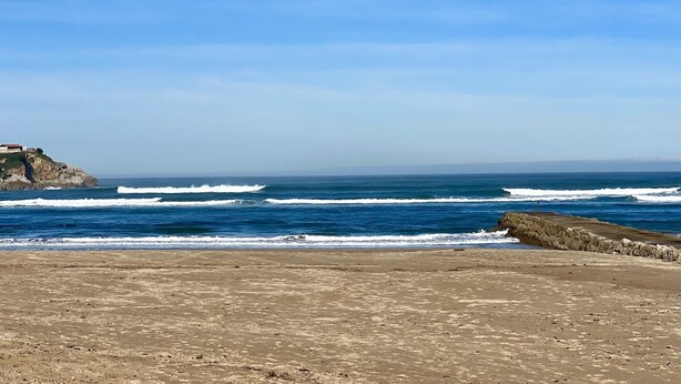 Una playa con olas rompiendo en la orilla, un acantilado rocoso con un edificio en la cima y un muro de piedra que separa la playa del acantilado.