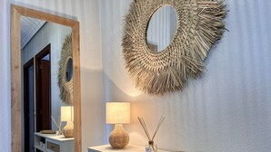 Un baño moderno con un espejo grande, un adorno circular tejido en la pared, un tocador blanco con una lámpara y una pequeña estantería con plantas.