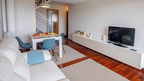 Un salon moderne avec un canapé blanc, une table à manger et des chaises, un téléviseur posé sur un meuble blanc, et un escalier menant à une autre pièce.