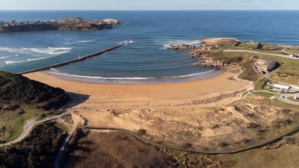 Vue aérienne d’une zone côtière avec une plage de sable, une digue rocheuse et un pont reliant la terre à la plage.
