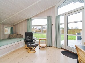 Une pièce avec un plafond en bois, un fauteuil en cuir noir, une petite table ronde, un banc avec des coussins et une grande porte-fenêtre coulissante donnant sur un espace extérieur.