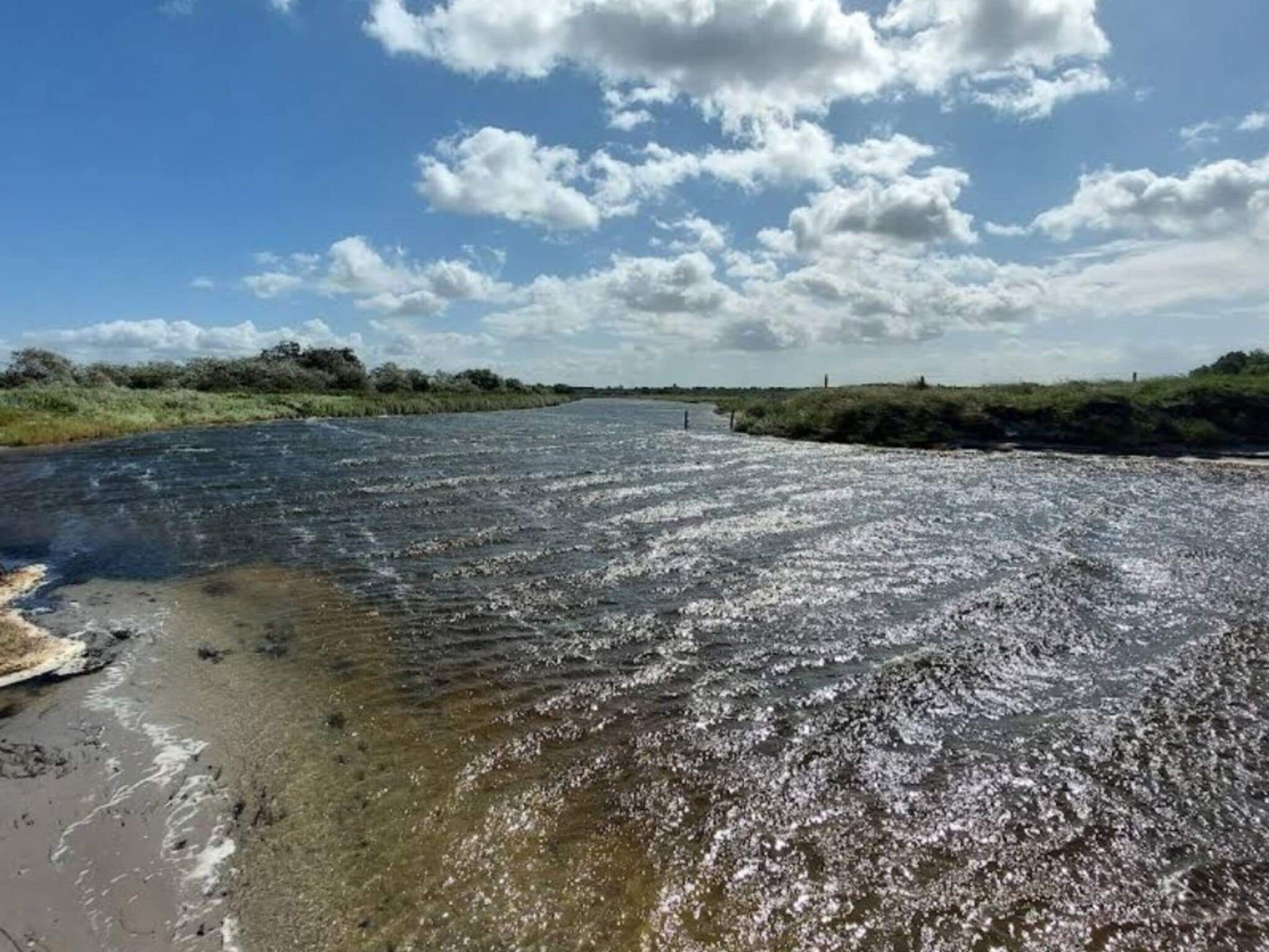 A river with clear water, surrounded by greenery and a partly cloudy sky.
