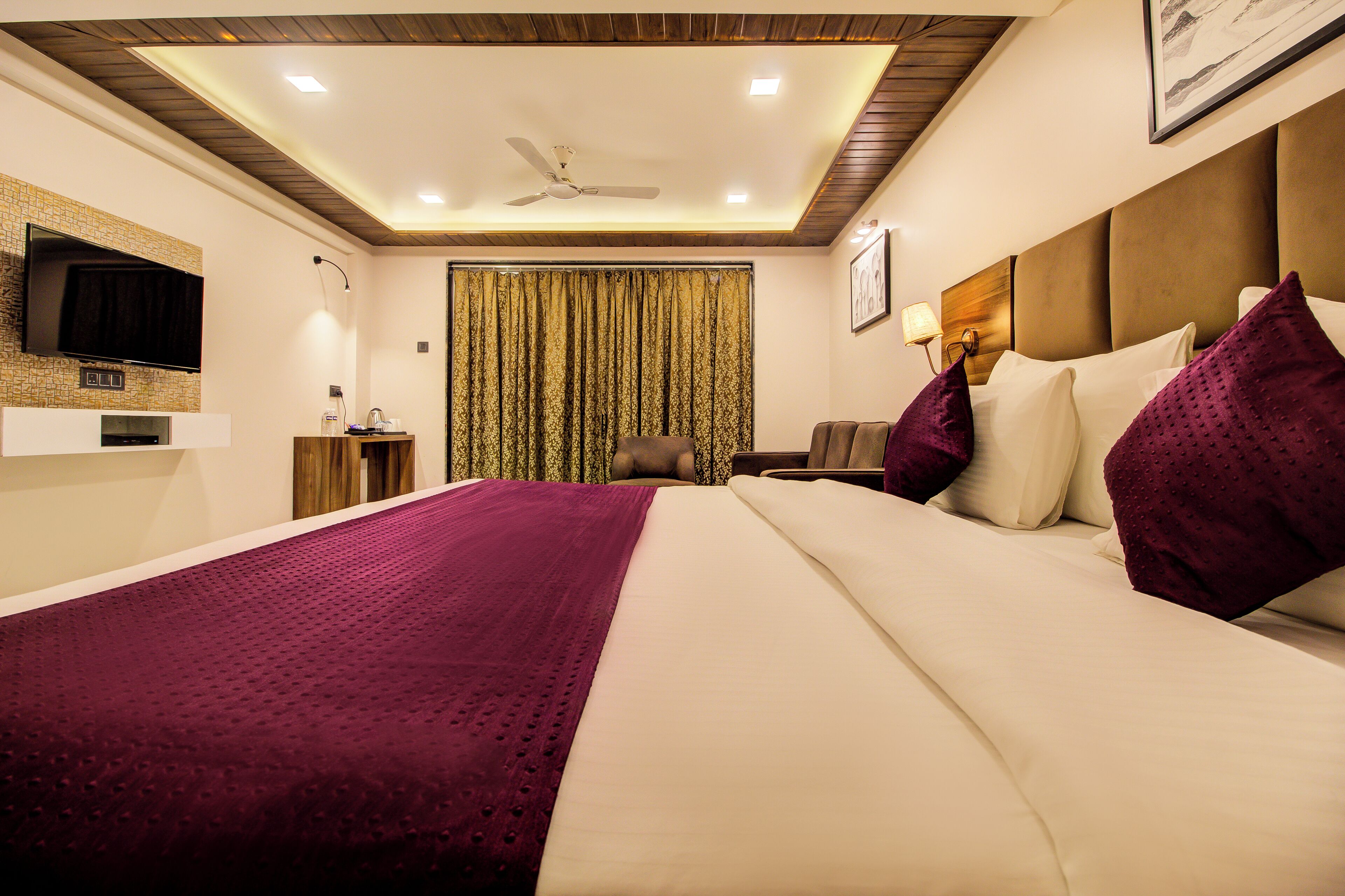 A hotel room with a large bed, a flat-screen TV mounted on the wall, a ceiling fan, and a wooden ceiling.