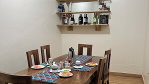 A dining area with a wooden table set for four, surrounded by chairs. Above the table, there are wooden shelves holding various bottles. - Rafiki Safari Lodge Bwindi (Bwindi)