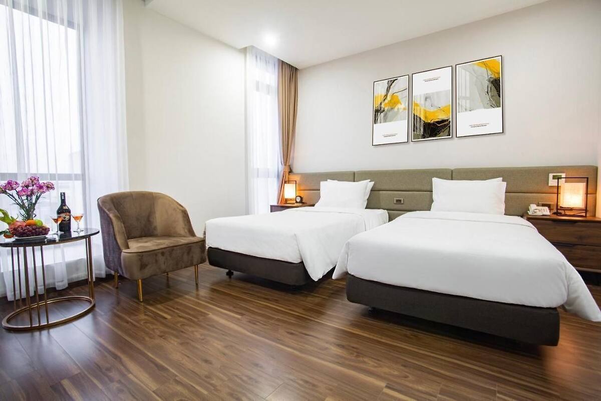 A hotel room with two beds, a chair, a small table with a fruit bowl, and artwork on the wall.