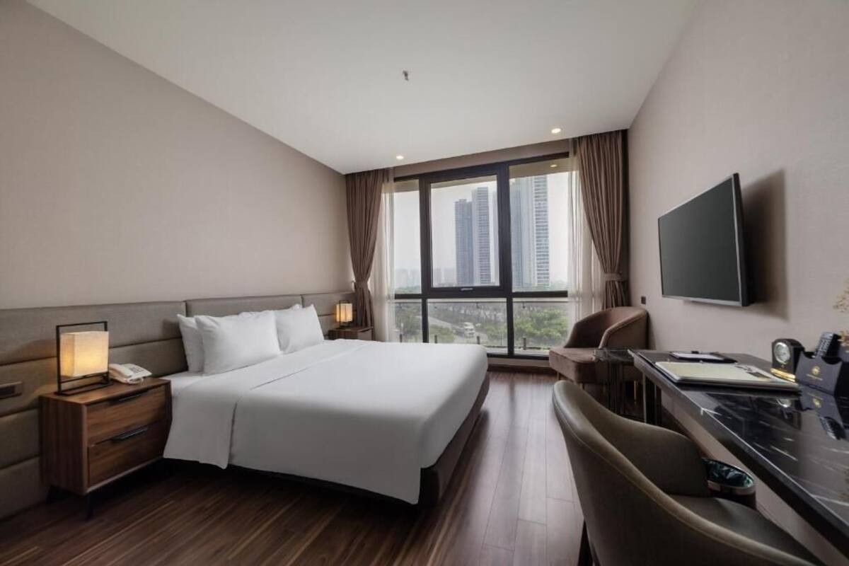 A modern hotel room with a large bed, a desk, a chair, a TV, and a city view.
