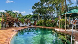 A clear swimming pool with lounge chairs and a covered seating area surrounded by lush greenery.