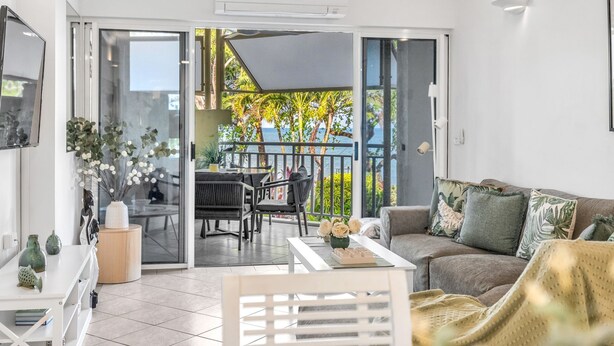 A modern living room with a sofa, a coffee table, and a dining area with chairs. There is a sliding glass door leading to a balcony with a view of greenery and a building.