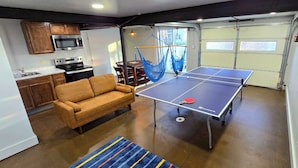 A compact living space with a ping pong table, a sofa, a hammock, and a kitchen area with wooden cabinets and a microwave. - The Green House! Sleeps 12 (Trenton)