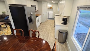 A modern kitchen with white cabinets, a stainless steel refrigerator, a coffee maker, and a view of the outdoors through large windows. - The Green House! Sleeps 12 (Trenton)