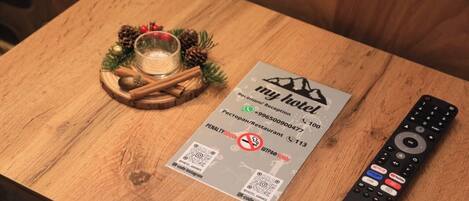 Una mesa de madera con una tarjeta de recepción de hotel, un mando a distancia y un vaso de agua con ramas de canela y piñas.