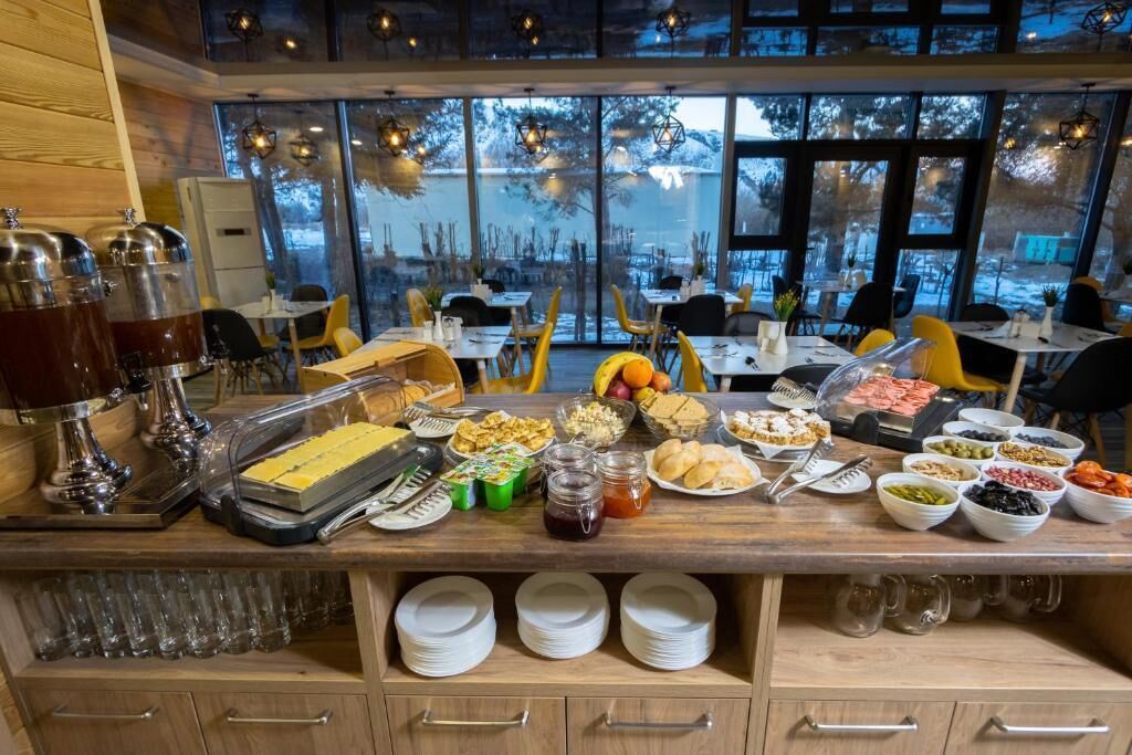 A buffet setup with various food items and condiments, wooden furniture, and a view of the outdoors through large windows.