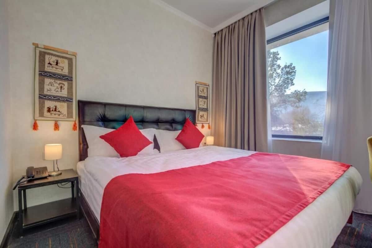Une chambre d’hôtel avec un grand lit, une literie rouge et blanche, une tête de lit agrémentée de coussins rouges, une table de chevet avec une lampe et une fenêtre avec des rideaux.