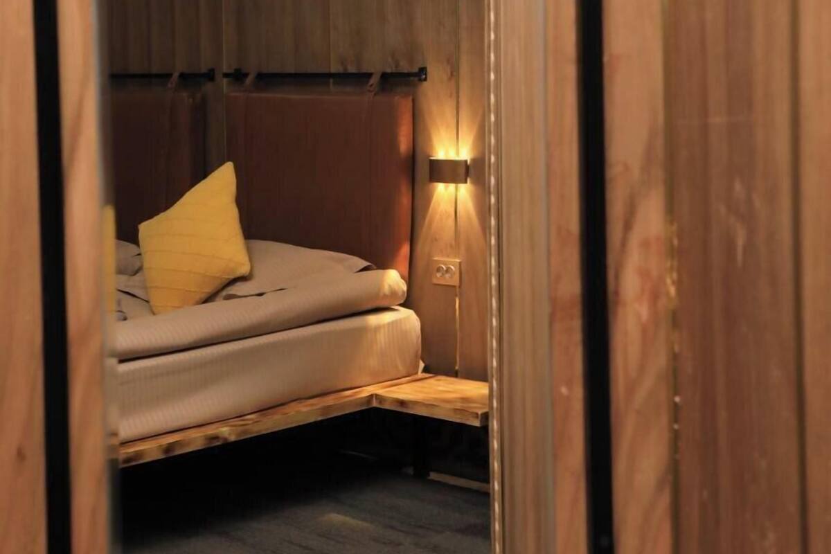 Une petite pièce comprenant un lit, une lampe fixée au mur et un petit banc.