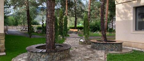 Un giardino curato con un vialetto in pietra, due aiuole circolari con alberi, una panchina e un edificio con una finestra.