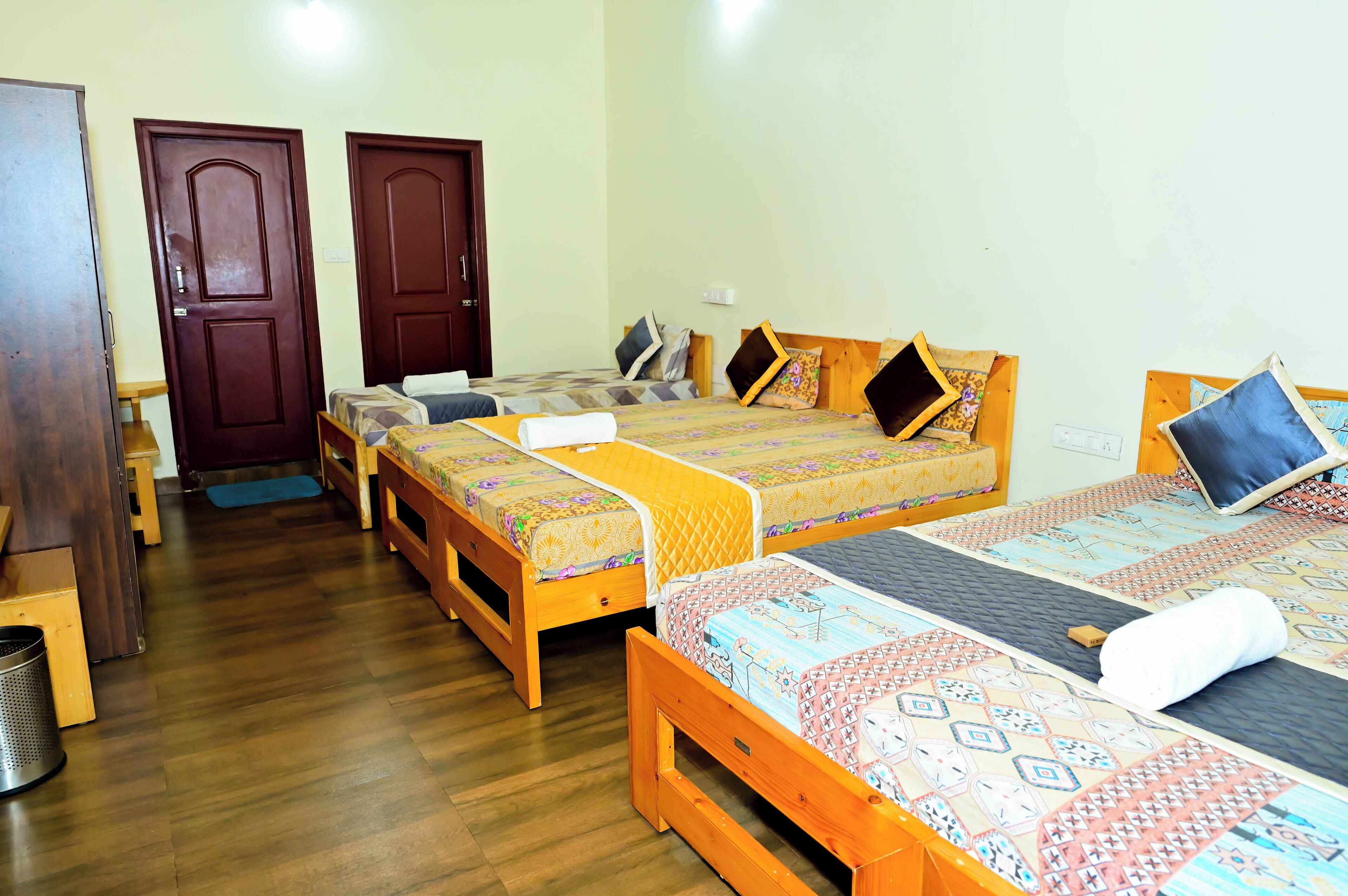 Una habitación con cuatro camas de madera, cada una con ropa de cama y almohadas estampadas. Hay dos puertas de madera y un banco de madera.