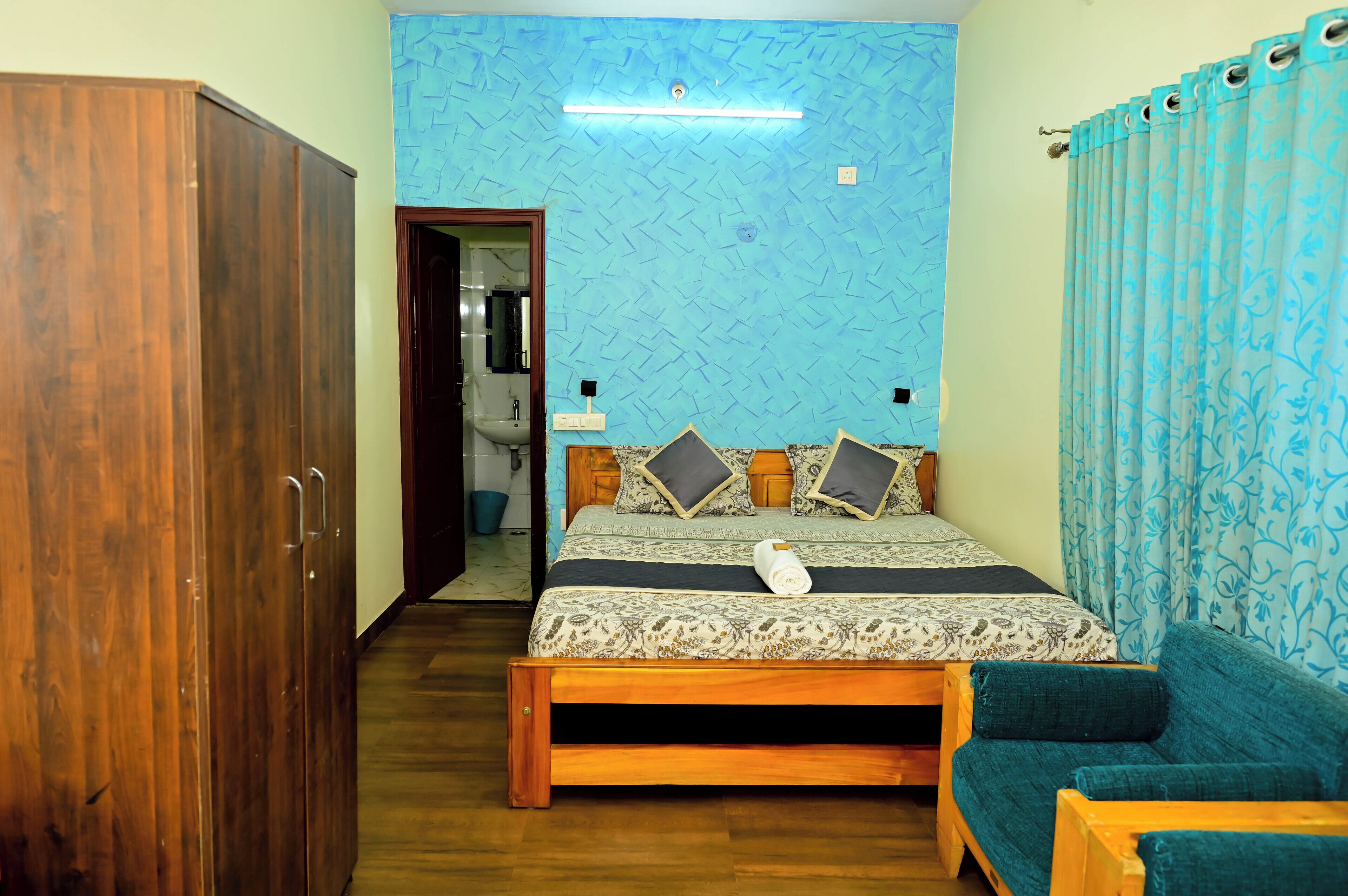 Un dormitorio con un armario de madera, una cama con ropa de cama estampada, un sofá azul y un baño visible a través de una puerta abierta.