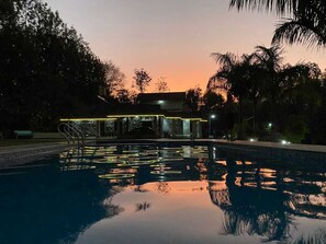 A swimming pool at dusk with reflections of lights, surrounded by trees and a building with illuminated railings. - Elegance Homestay (Chikkamagaluru)