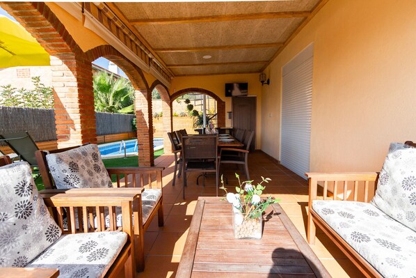 Un patio al aire libre con mobiliario de madera, una mesa de comedor y una piscina al fondo.
