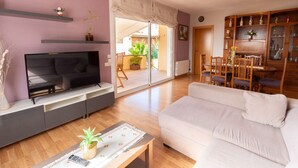 Una sala de estar con televisor de pantalla plana, un sofá, una mesa de centro con una planta y una zona de comedor con muebles de madera.