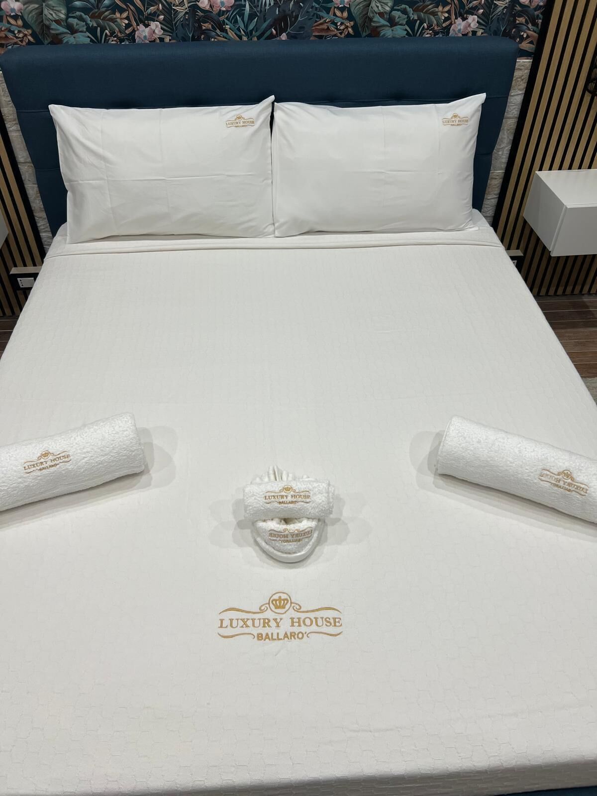 Ein ordentlich bezogenes Bett mit weißen Leinen und Kissen, einem Kopfteil sowie zwei Handtüchern mit dem Schriftzug 'Luxury House Ballaro'.