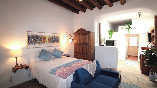 Un dormitorio con cama, mesita de noche, lámpara y un sillón azul. Hay un armario de madera y un cuadro en la pared.