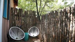 Una valla de madera, dos sillas blancas y un bote de basura verde en un área exterior sombreada.