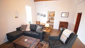 Una sala de estar con un sofá seccional, un sillón individual, una mesa de centro y una mesita con una lámpara. Hay una alfombra en el suelo, y la habitación tiene paredes blancas con una viga de techo de madera.