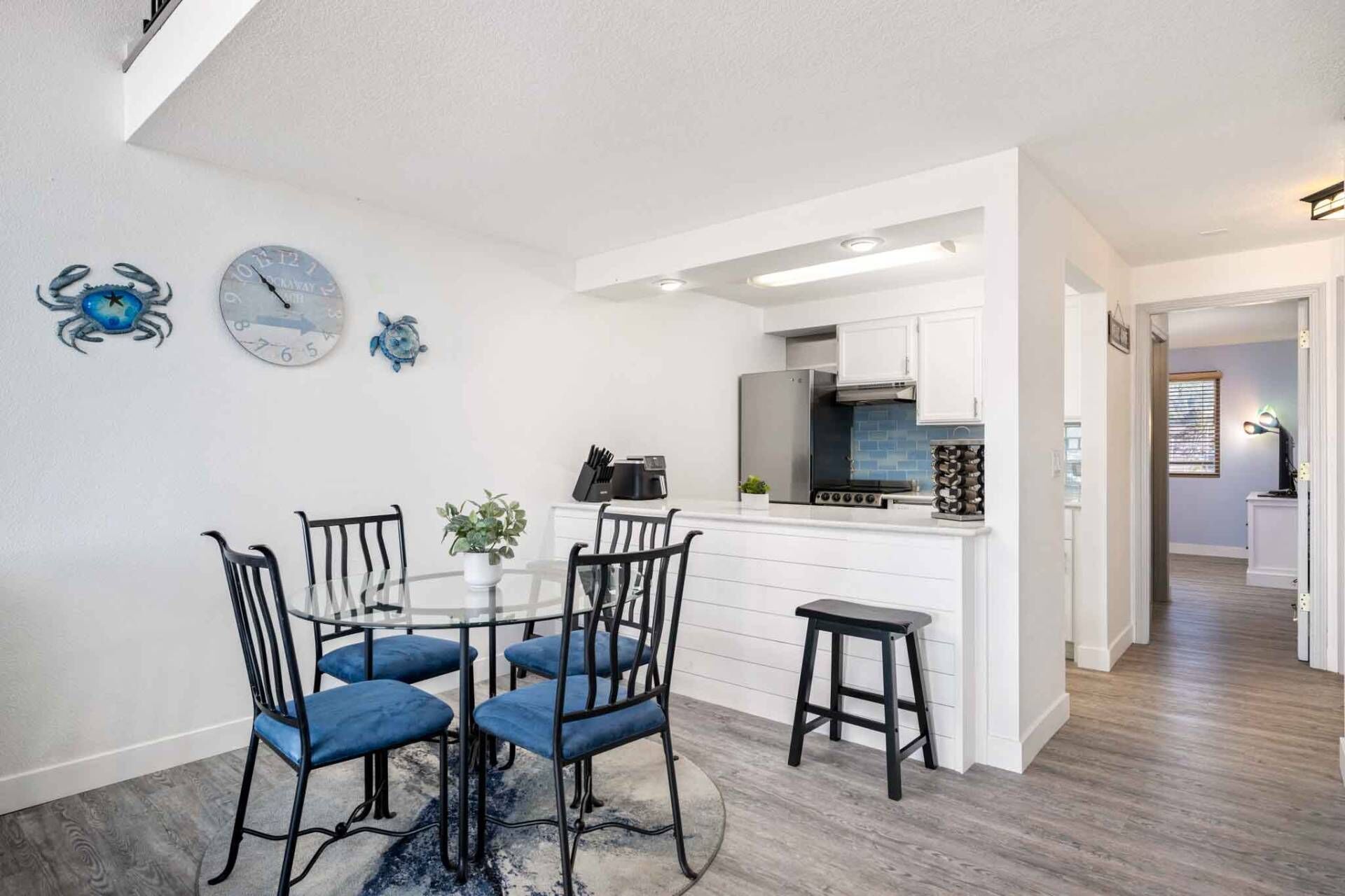 A modern kitchen with white cabinets, a stainless steel refrigerator, and a built-in oven. There is a dining area with a round table and four blue chairs. Decorative wall clocks and a turtle decoration are visible.