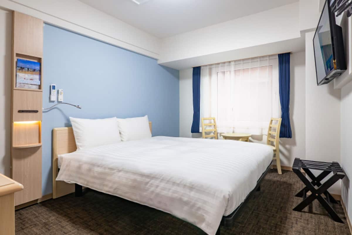 A hotel room with a large bed, a desk with a chair, a TV mounted on the wall, and a window with curtains.