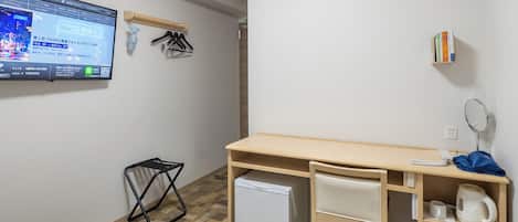 A hotel room with a bed, a desk, a small refrigerator, a wall-mounted TV, an air conditioner, a bookshelf, and a waste bin.