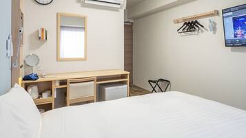 A hotel room with a bed, a desk with a mirror, a small kitchenette, an air conditioning unit, a TV, and a wall-mounted clock.