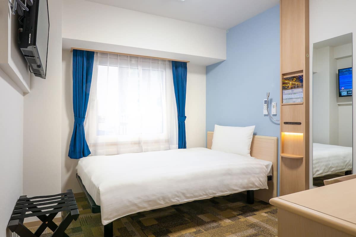 A hotel room with a bed, a desk, a chair, a window with curtains, a mirror, and a wall-mounted TV.